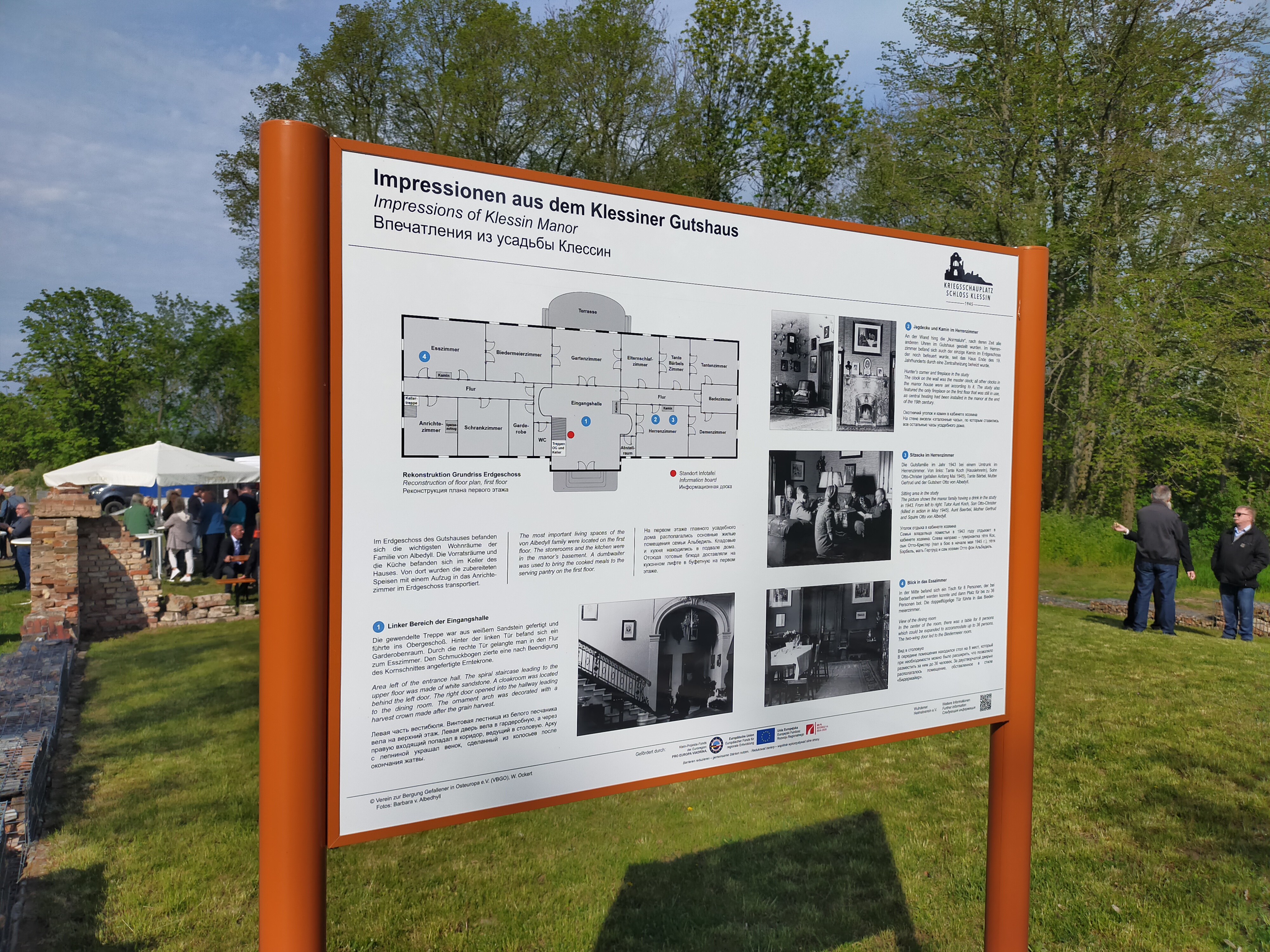 Der Blick fällt auf eine Informationstafel, die inmitten einer Grasfläche zwischen Grundmauern steht. Die Überschrift der Tafel lautet "Impressionen aus dem Klessiner Gutshaus". Darauf sind ein Grundriss, historische Fotos sowie Texte abgebildet