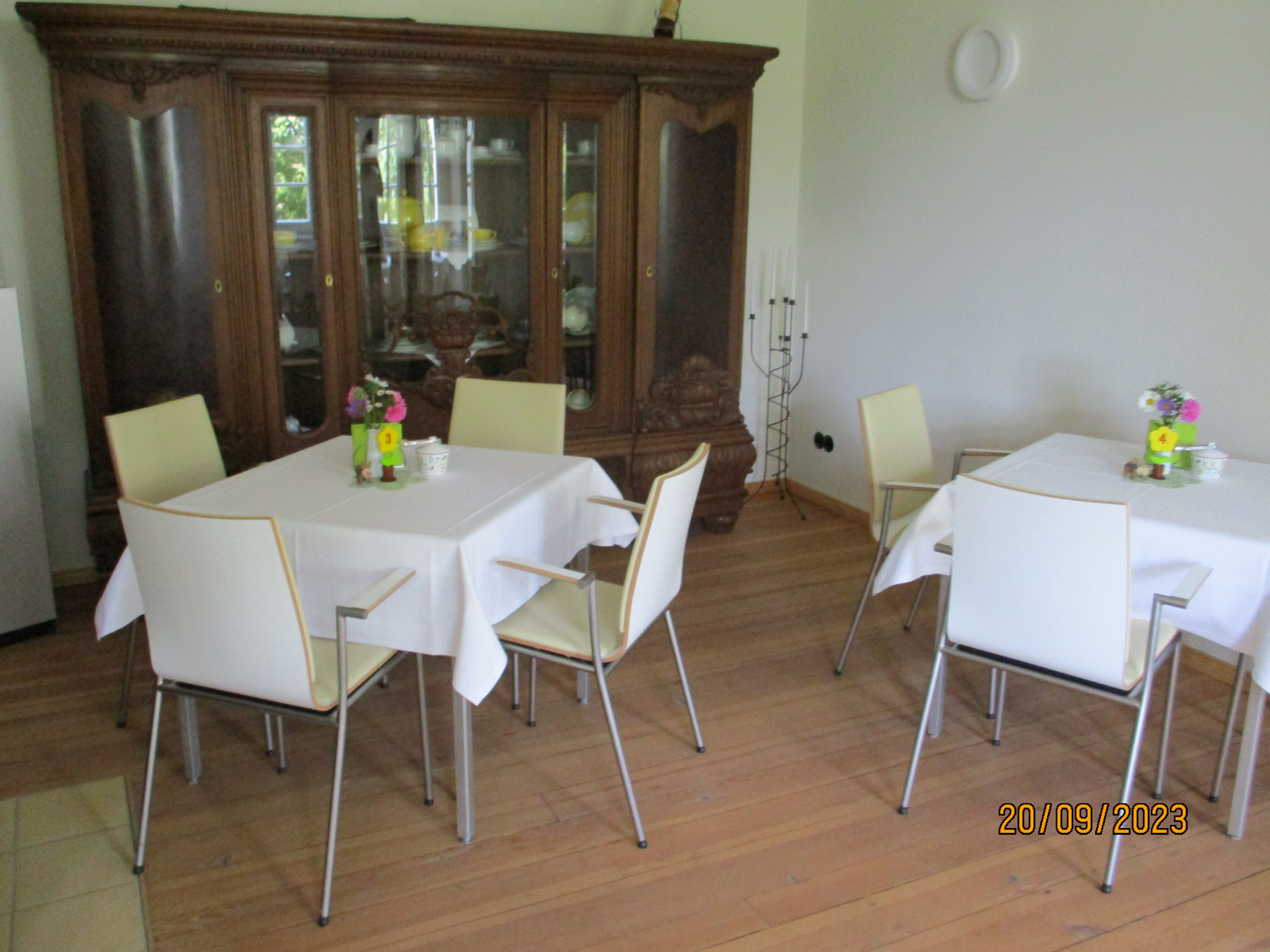 Ein Raum mit Parkettboden, einem historisierenden Holzschrank und zwei mit weißen Tischdecken bezogene Tische, die mit Blumensträußen und Zuckerdosen ausgestattet sind, laden zum Kaffeetrinken ein