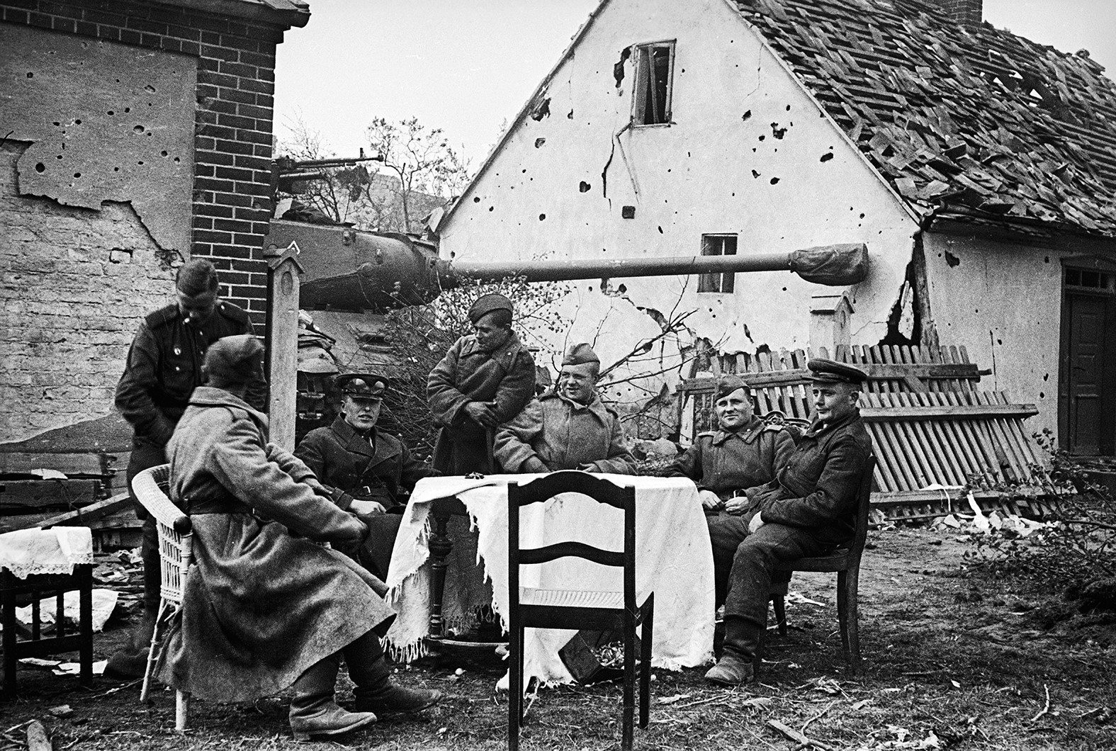 Das Foto zeigt im Vordergrund eine Gruppe sowjetischer Soldaten, die unter unter freiem Himmel um einen Tisch sitzt .   Im Hintergrund erkennt man einen offensichtlich abgestellten Kampfpanzer zwischen zwei durch kriegsspurengezeichneten Häusern. 