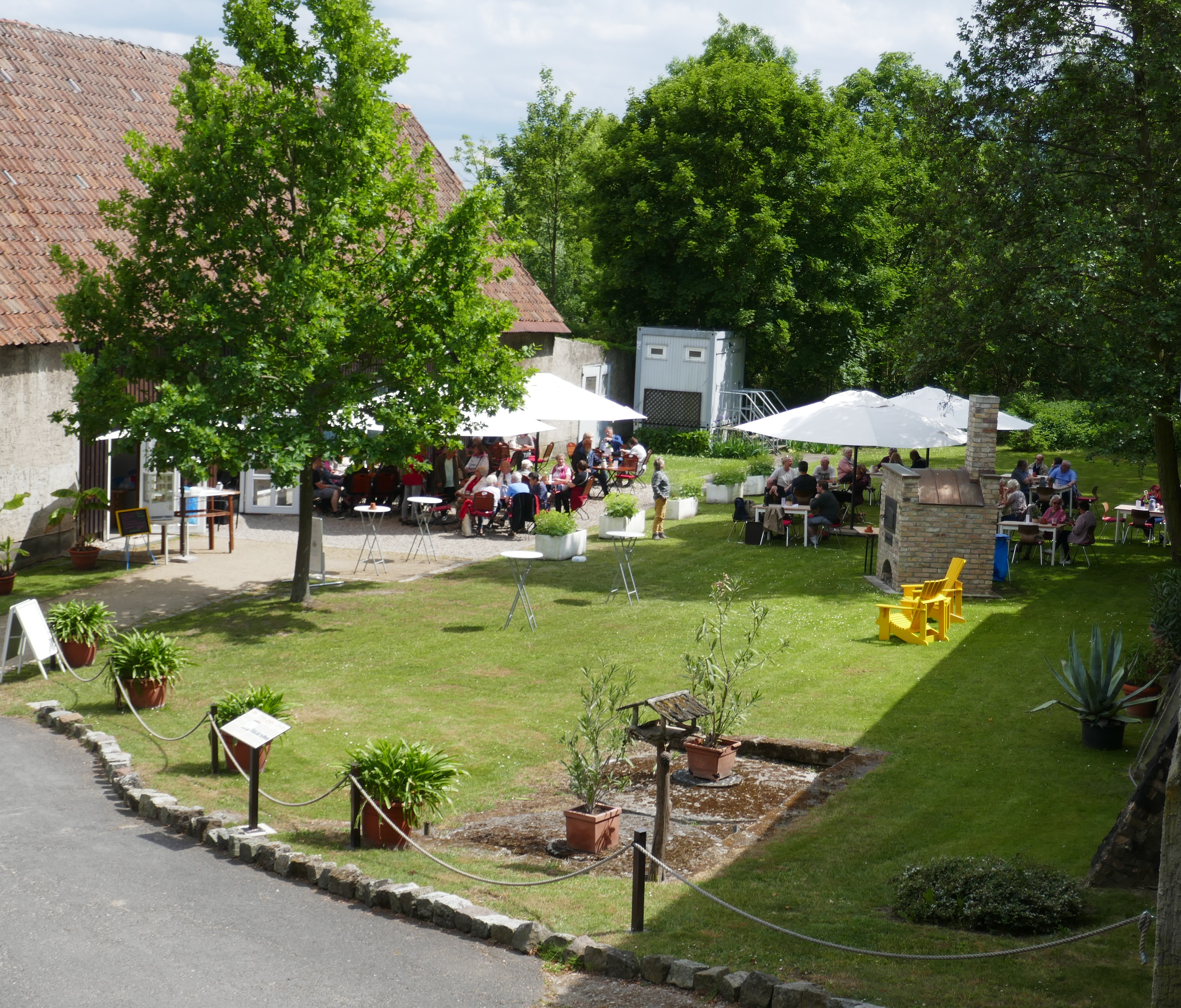 Blick in einen gepflegten und sonnenbeschienenen Garten, in dem zahlreiche Besucher an Tischen verweilen und unter aufgespannten Sonnenschirmen Schatten suchen
